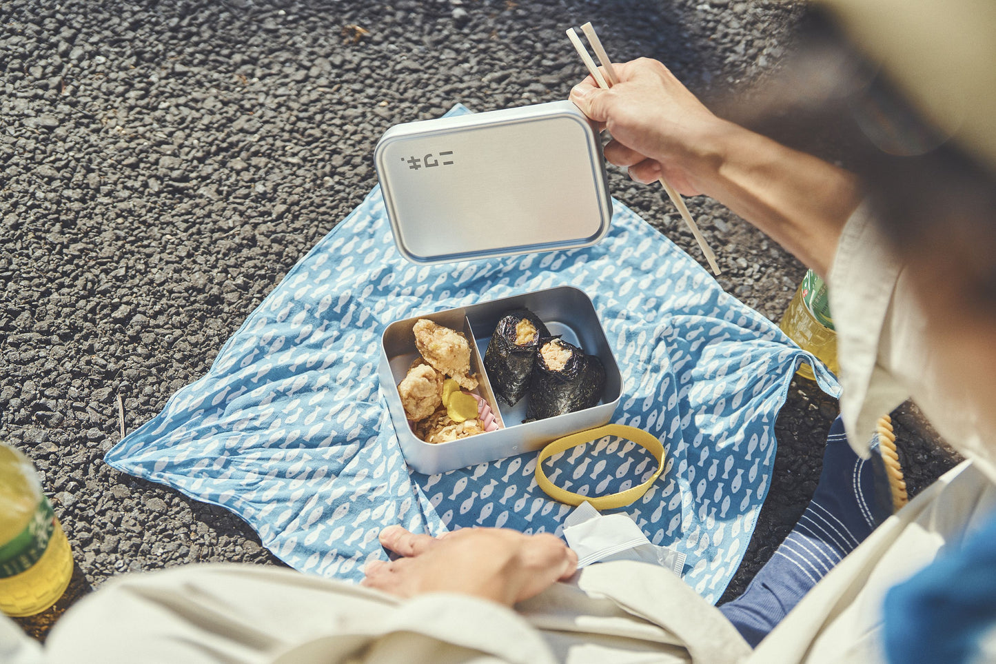 Person sitting on a blue patterned blanket with a lunch box and food items outdoors.