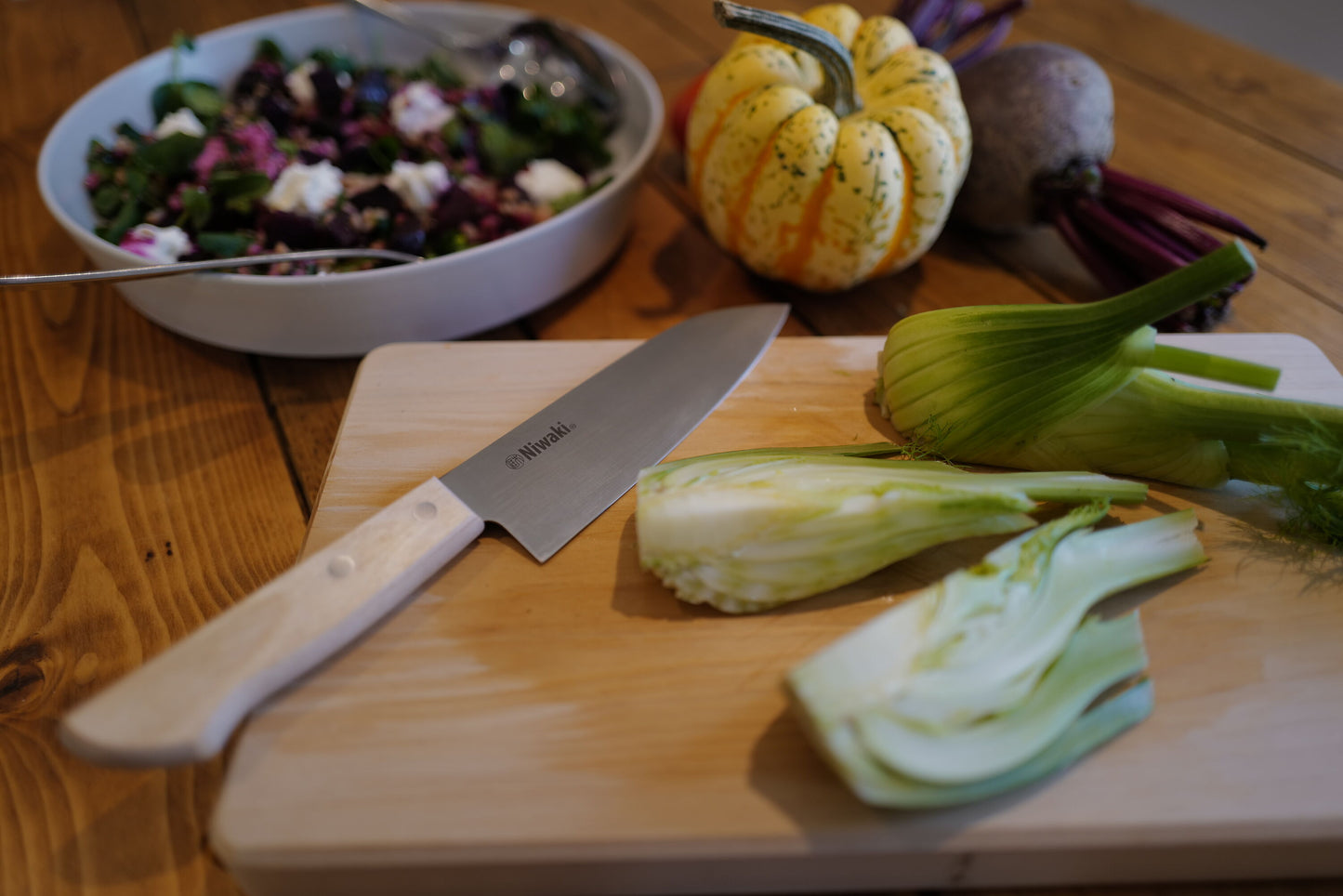 Sliced zucchini on a wooden cutting board with a knife and a bowl of salad in the background.