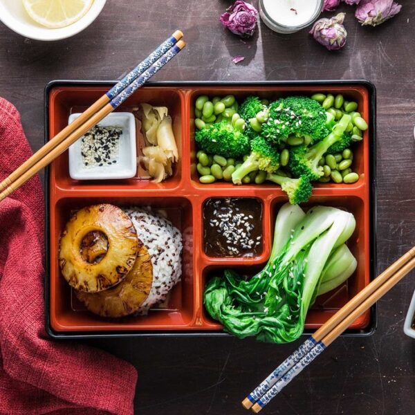 A black compartment bento box filled with a meal: broccoli with edamame, bok choy, rice with grilled pineapple, and small dipping dishes. Chopsticks rest on the box and two sides, and the scene is styled on a dark tabletop with a cloth napkin, sauce, and flowers.
