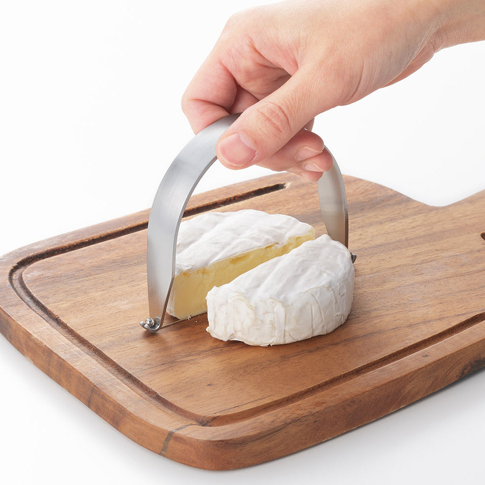 A hand using a curved stainless steel cheese slicer on a round wheel of brie cheese resting on a wooden cutting board, with a green 'New' badge in the top-left corner.