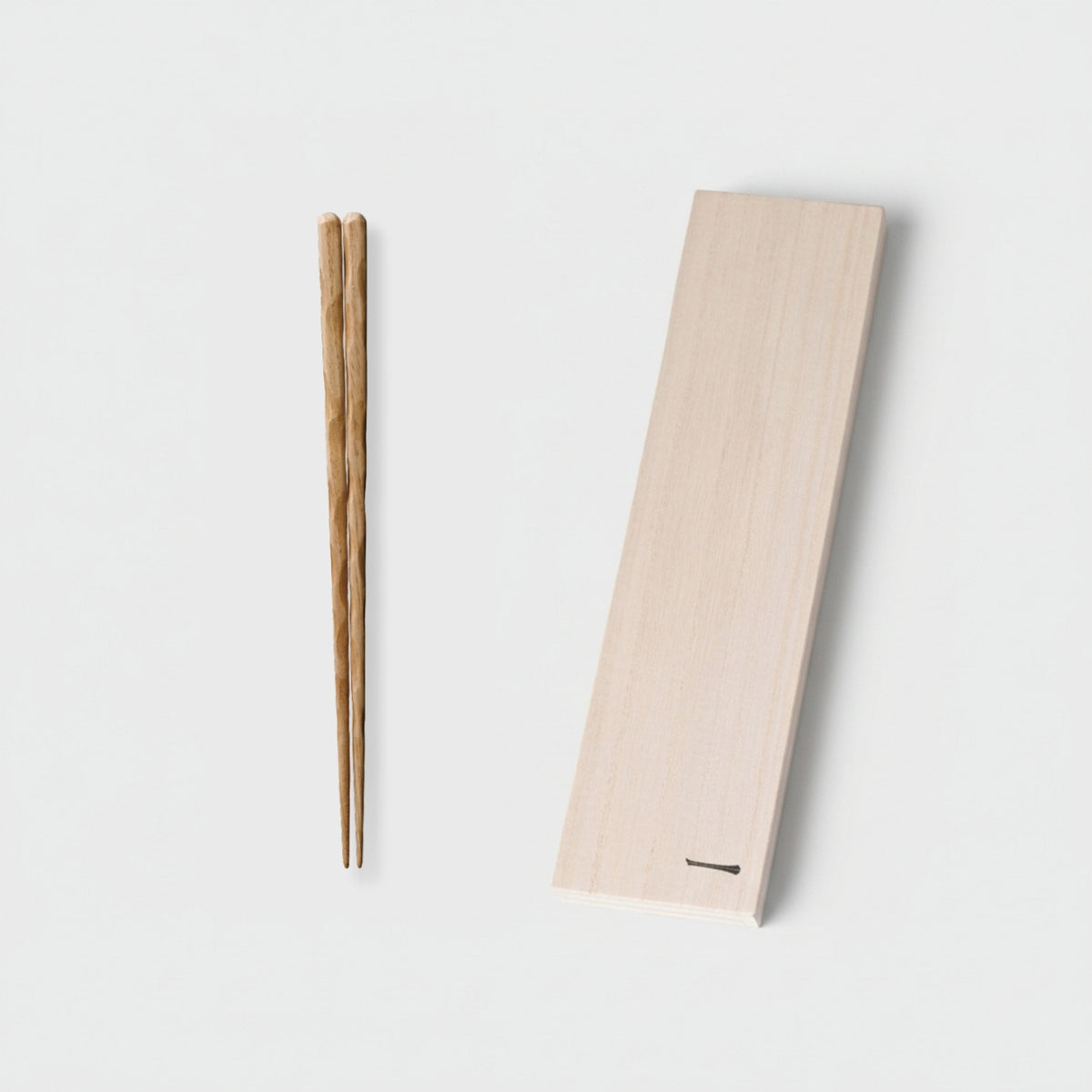 A pair of light brown wooden chopsticks placed beside a pale natural wood rectangular holder with a small slot at the bottom, photographed on a clean white background.