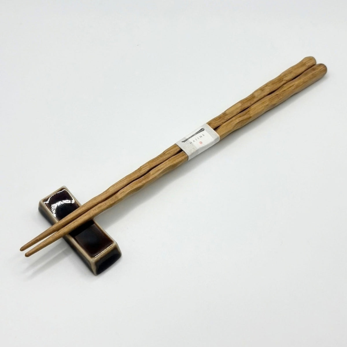 A pair of natural wood chopsticks resting on a small black ceramic chopstick holder, with a white paper band wrapped around the chopsticks, set against a plain white background.