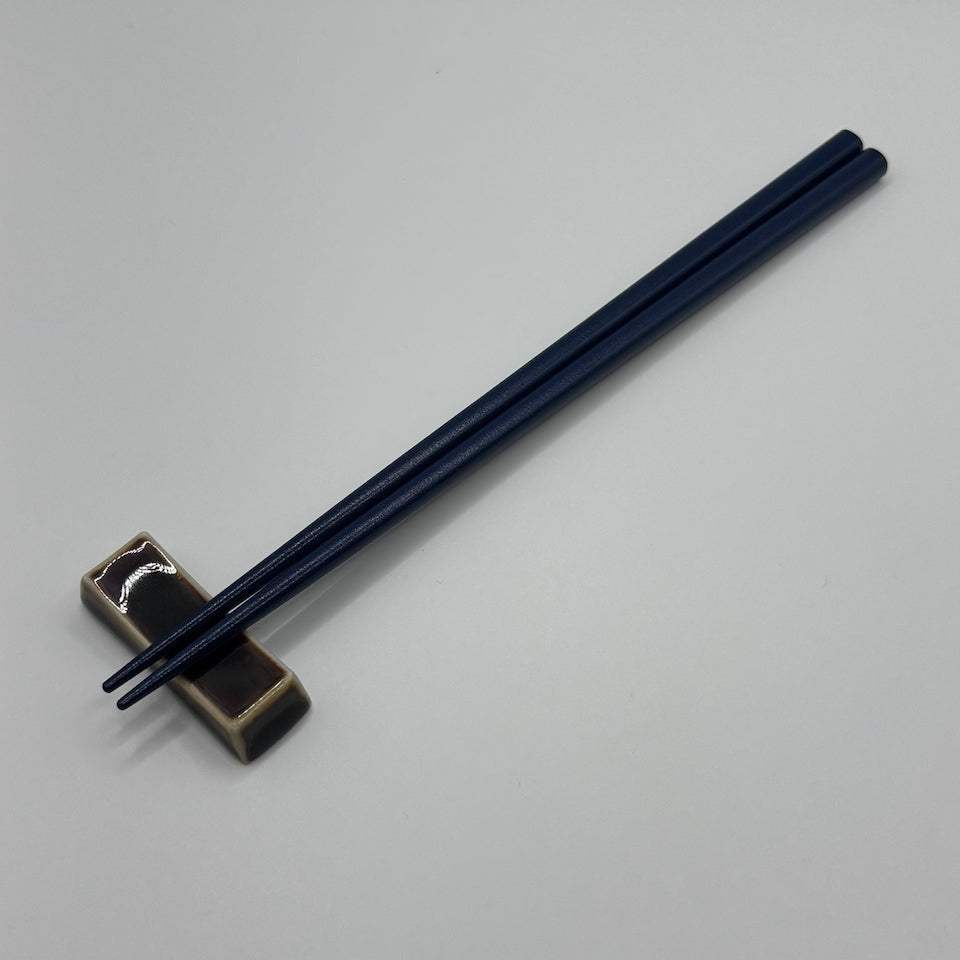 A pair of dark navy blue chopsticks resting on a small rectangular metallic holder against a plain light gray background.