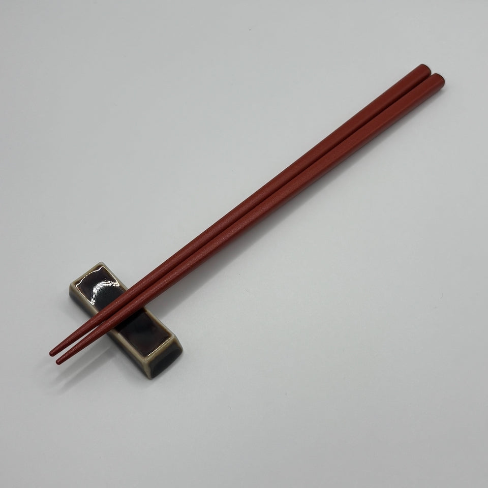 A pair of dark reddish-brown wooden chopsticks resting on a small black and beige ceramic chopstick holder, set against a plain light gray background.