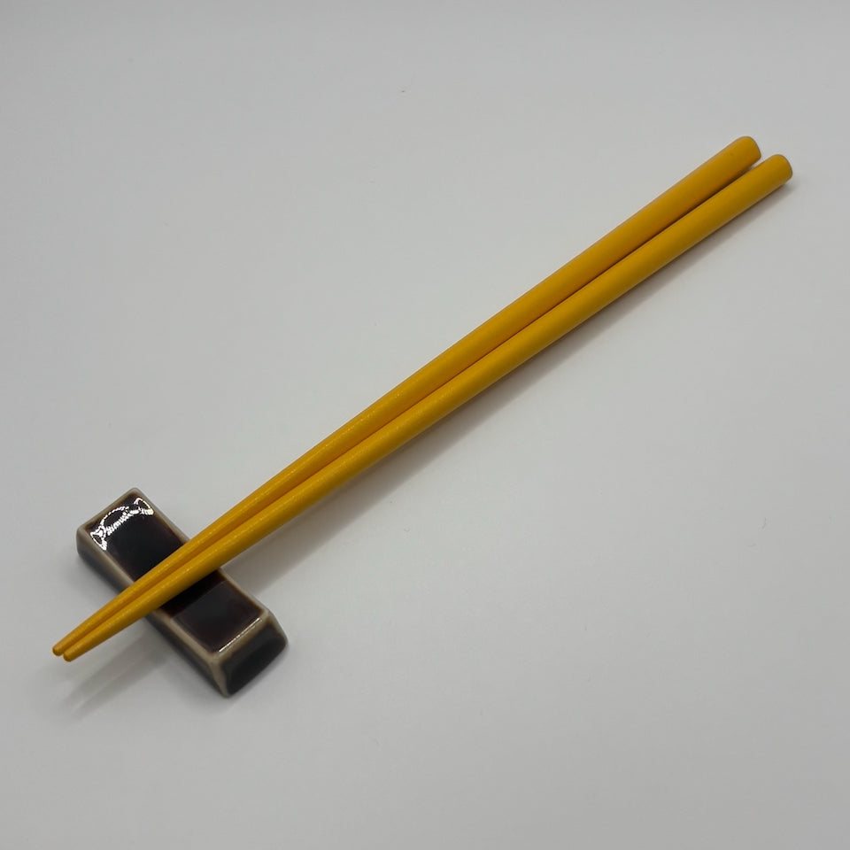 A pair of yellow wooden chopsticks resting on a small dark ceramic chopstick holder against a plain light gray background.