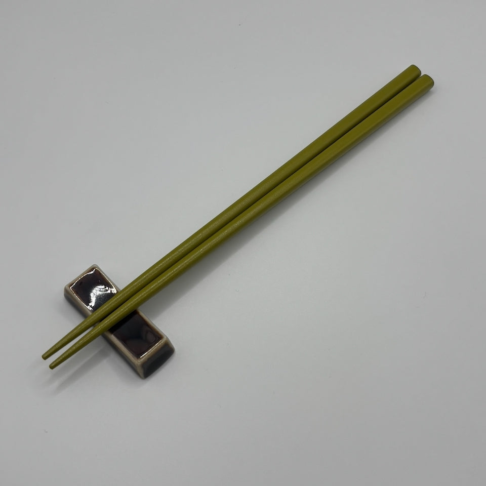 A pair of olive green chopsticks resting on a small black and silver chopstick holder against a plain light gray background.