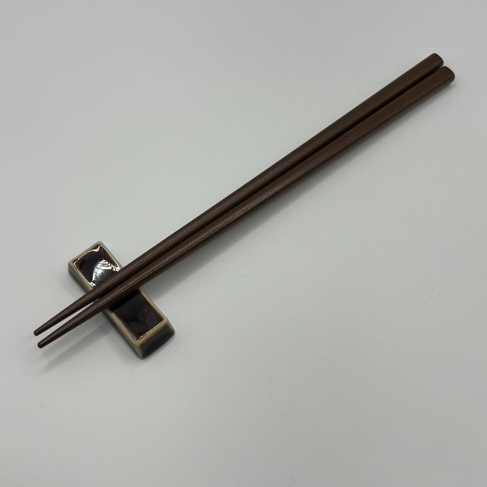 A pair of dark brown wooden chopsticks resting on a small black and beige ceramic chopstick holder against a plain light gray background.