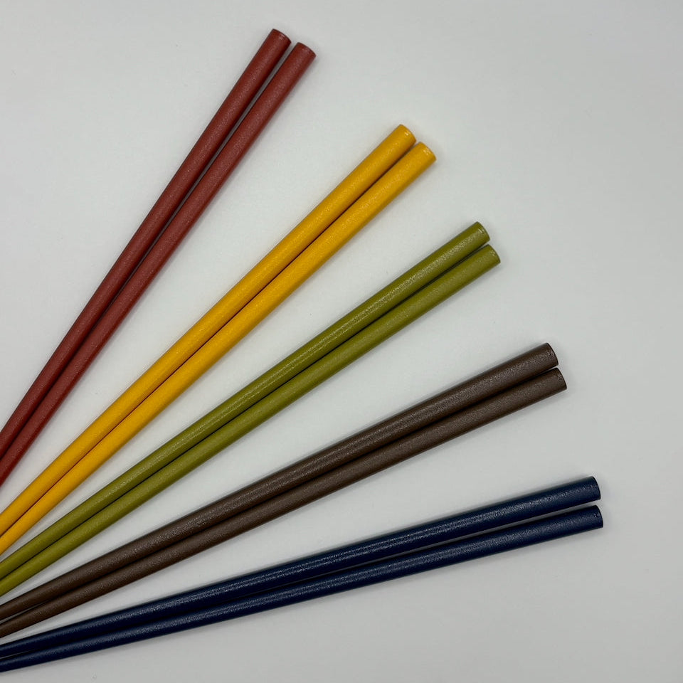 Six pairs of smooth, polished wooden chopsticks in different colors (red, yellow, olive green, dark brown, and navy blue) arranged in a fan shape on a plain white background.