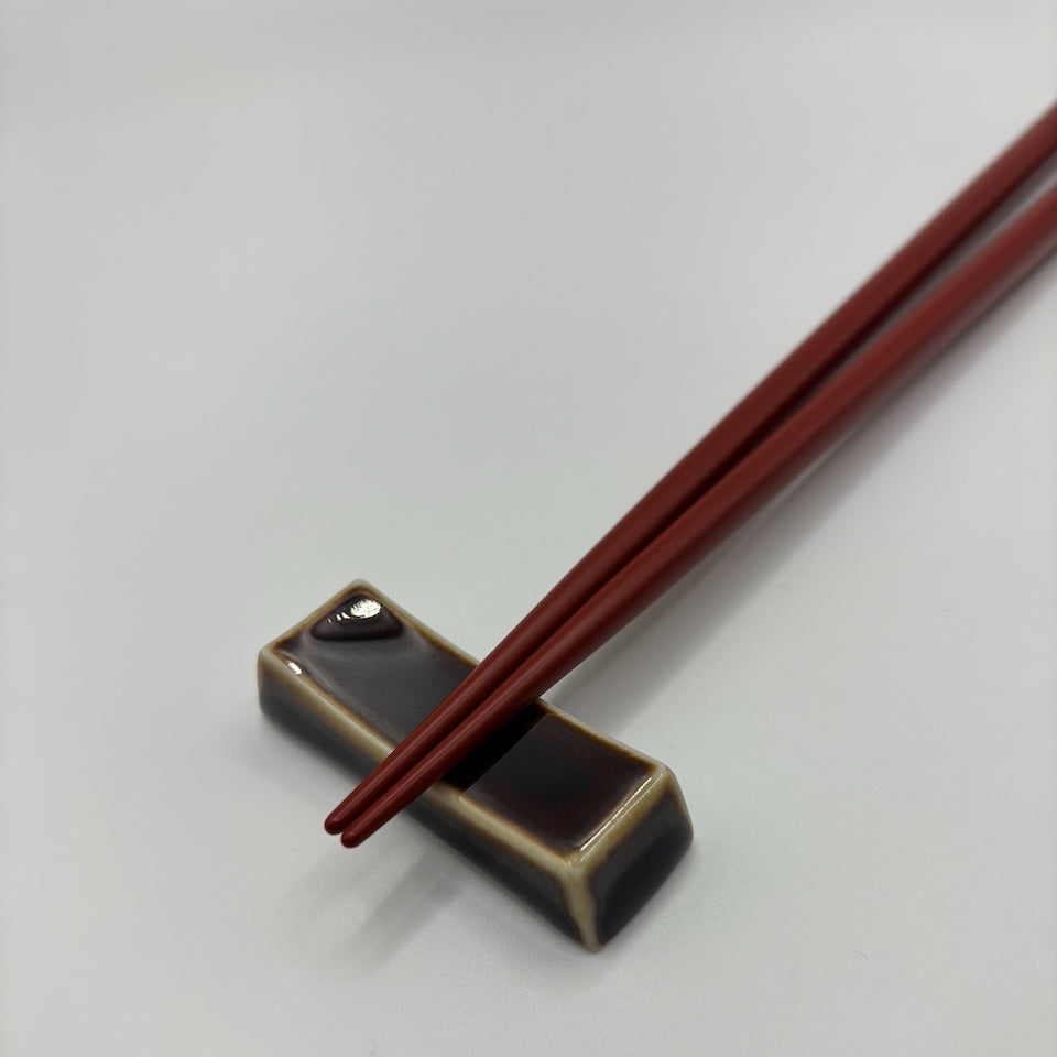 A pair of dark reddish-brown wooden chopsticks resting on a small rectangular black and beige ceramic chopstick holder against a plain light gray background.