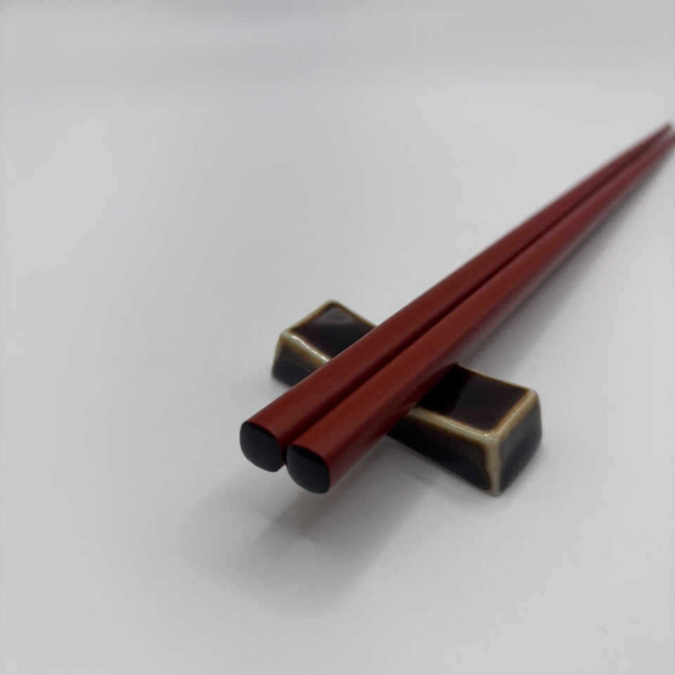 A pair of dark red wooden chopsticks resting on a black and gold square chopstick holder against a plain light gray background.
