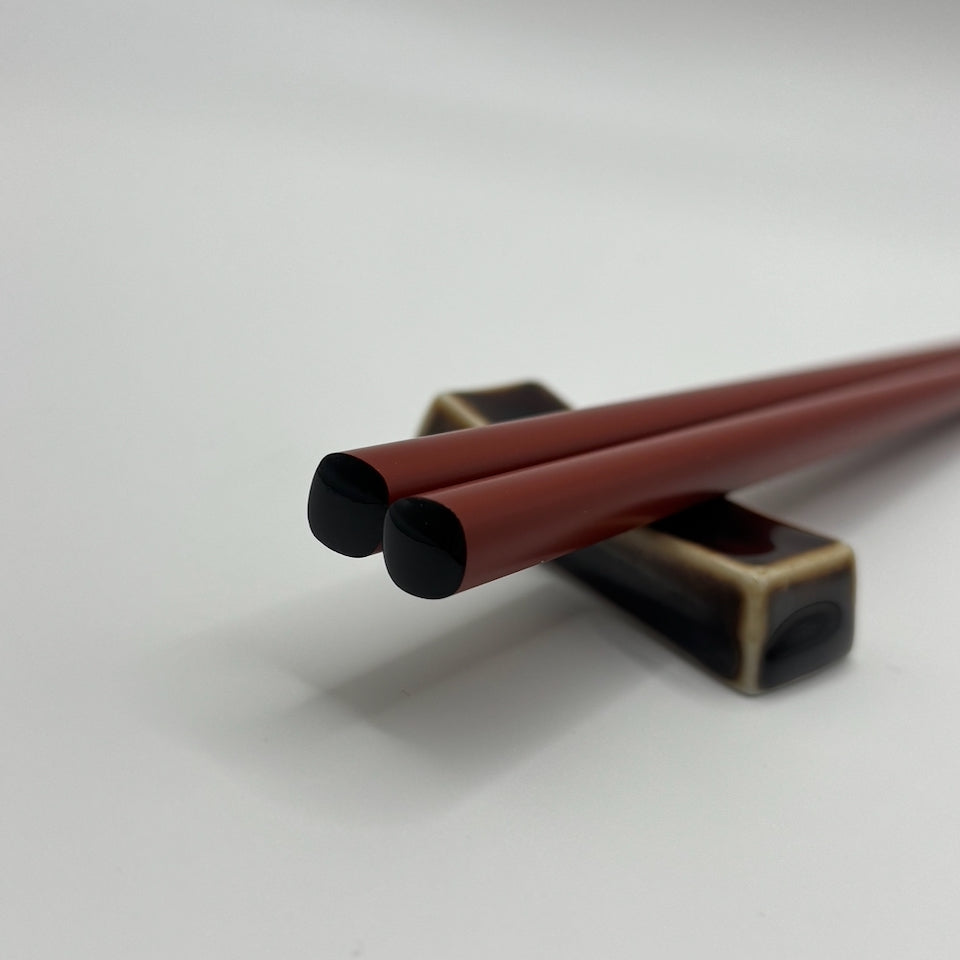 A pair of dark red lacquered chopsticks resting on a black and gold chopstick holder against a clean white background.