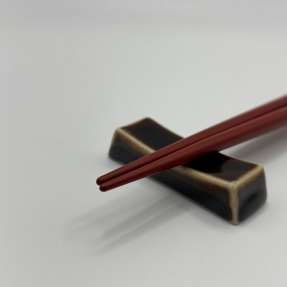Red wooden chopsticks resting on a dark brown and beige ceramic chopstick holder against a plain light gray background.