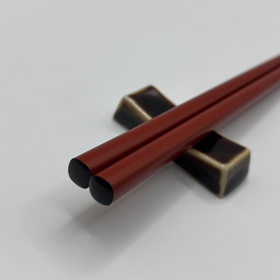 A pair of dark red chopsticks resting on a black and gold ceramic chopstick holder against a light gray background.