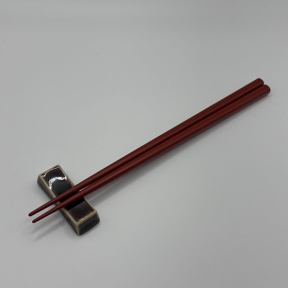 A pair of dark reddish-brown wooden chopsticks resting on a small black and beige ceramic chopstick holder against a neutral gray background.