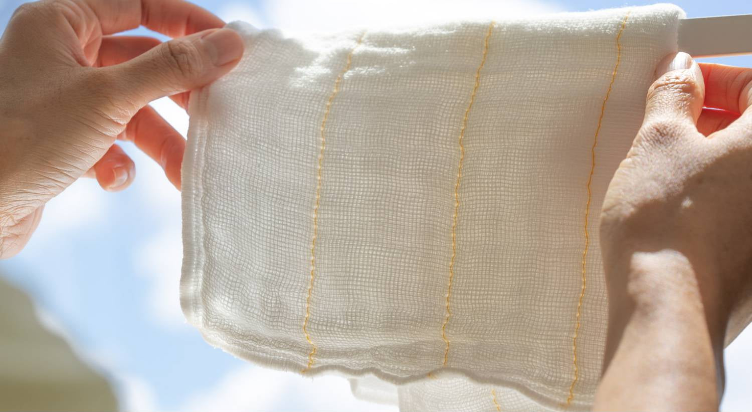Hands holding a piece of white fabric with yellow stitching against a blue sky.