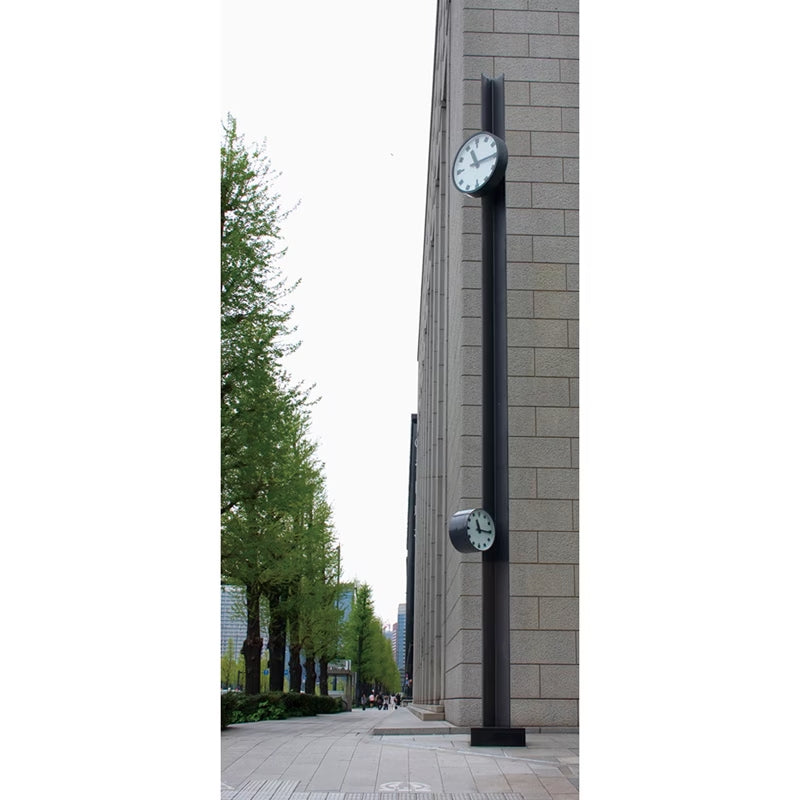 A tall black outdoor clock post mounted on a gray stone building, with a second smaller clock attached lower down. A sidewalk and green trees are visible along the left side of the scene.