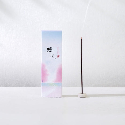 A minimalist incense holder setup featuring a tall rectangular box with a pastel blue and pink gradient design and black calligraphy, next to a single burning incense stick in a white holder, set against a white wall with soft leaf shadows.