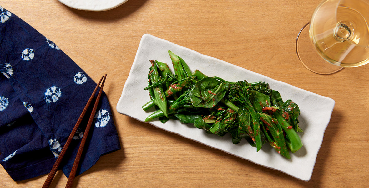 Plated green vegetables on a white rectangular plate with chopsticks and a blue napkin on a wooden table.