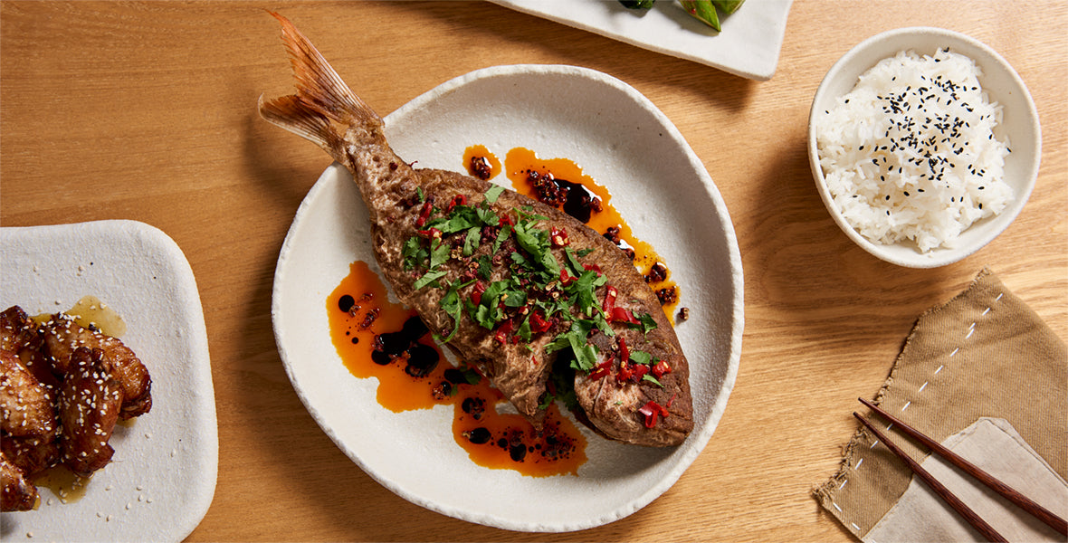 Steamed fish with garnishes on a white plate, served with rice and chopsticks on a wooden table.